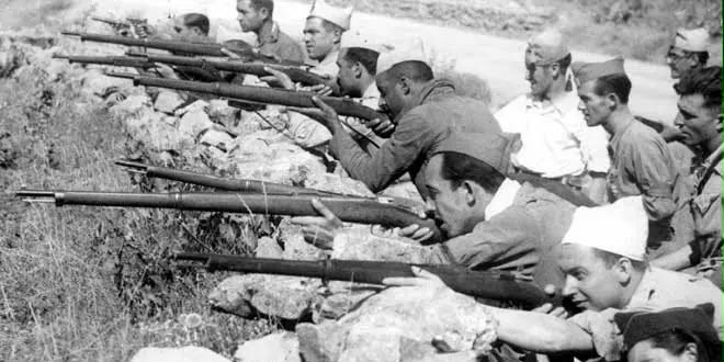 Fotografía en blanco y negro de soldados apostados tras un muro de piedra con sus fusiles durante la Guerra Civil.